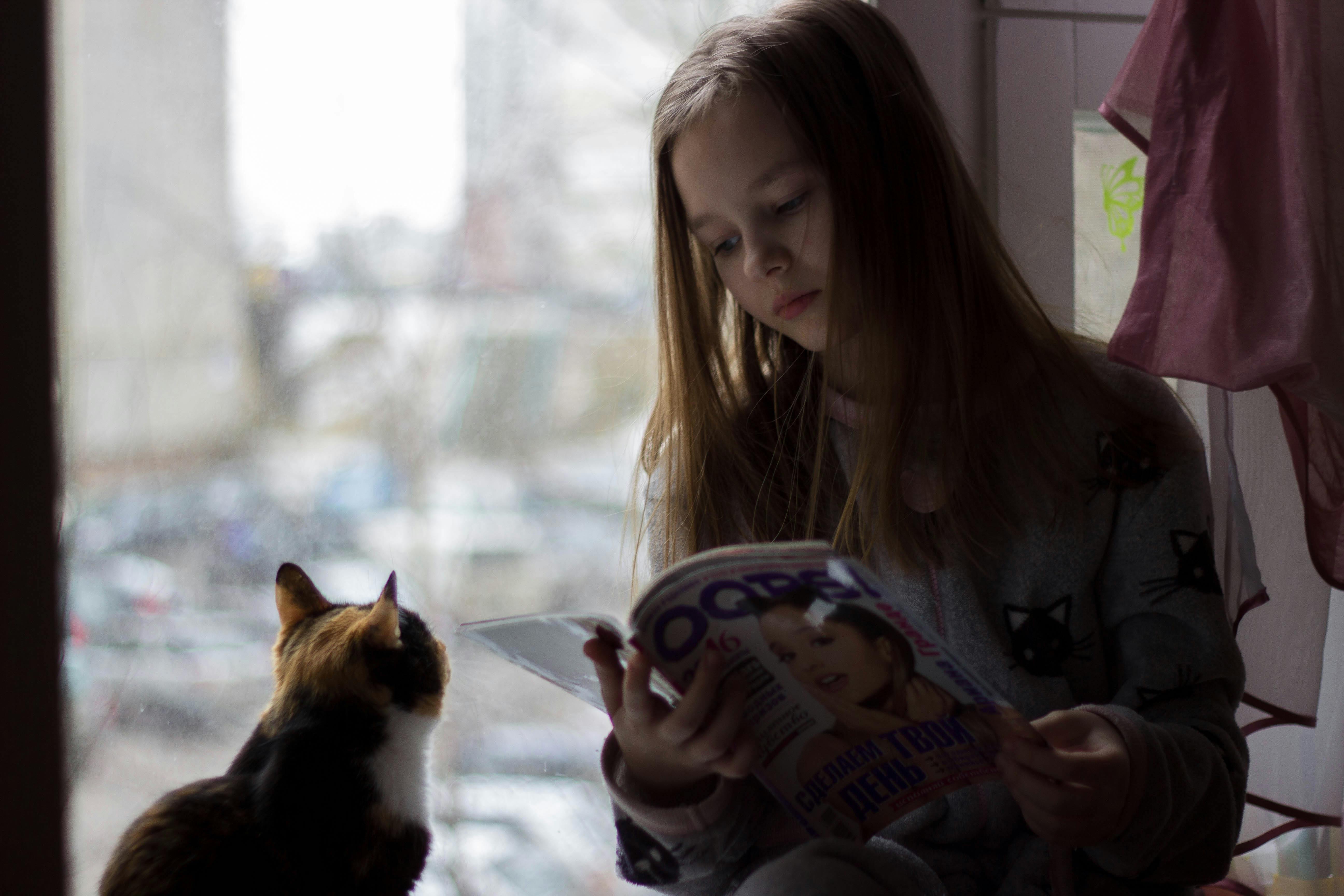 serious girl reading magazine sitting with quiet cat on windowsill