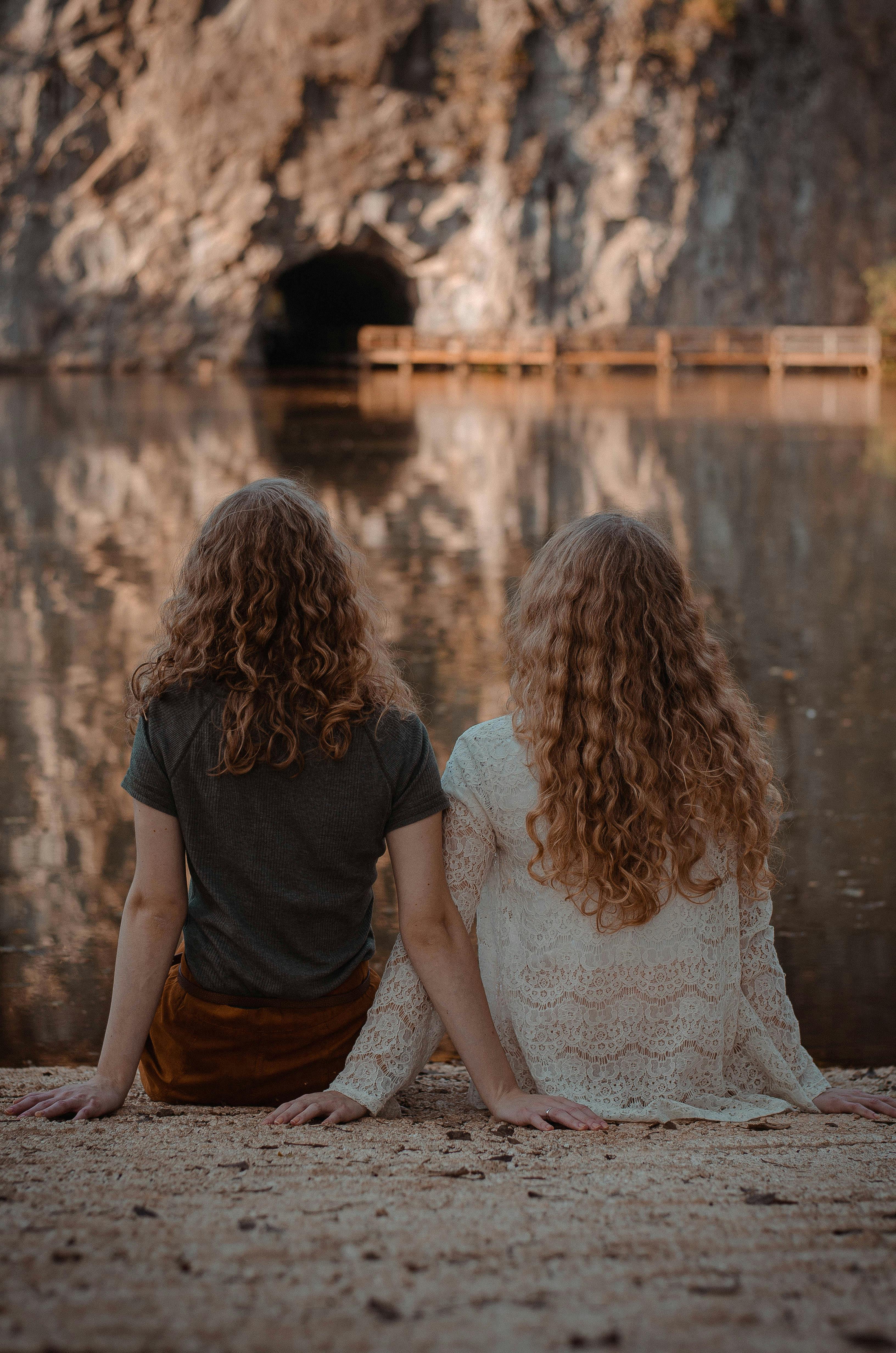 photography of two women sitting on ground facing on body of water