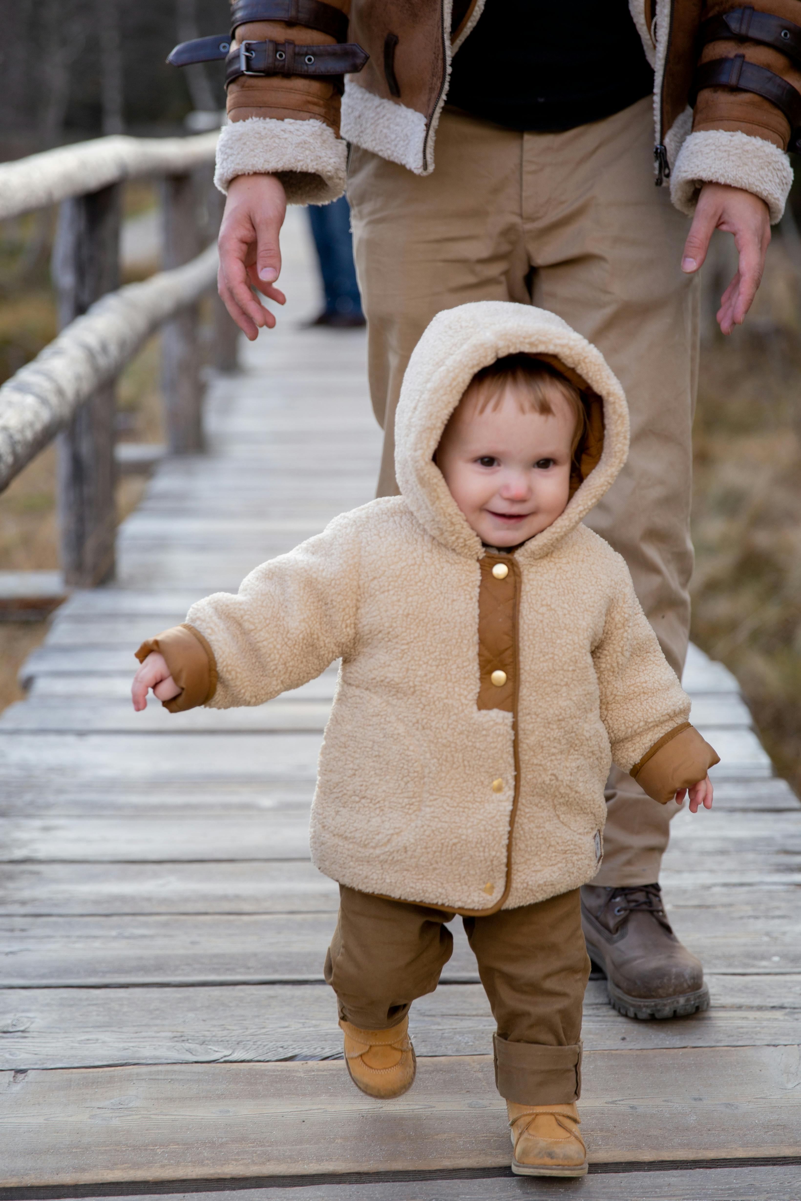 child in brown jacket and brown pants walking on wooden bridge