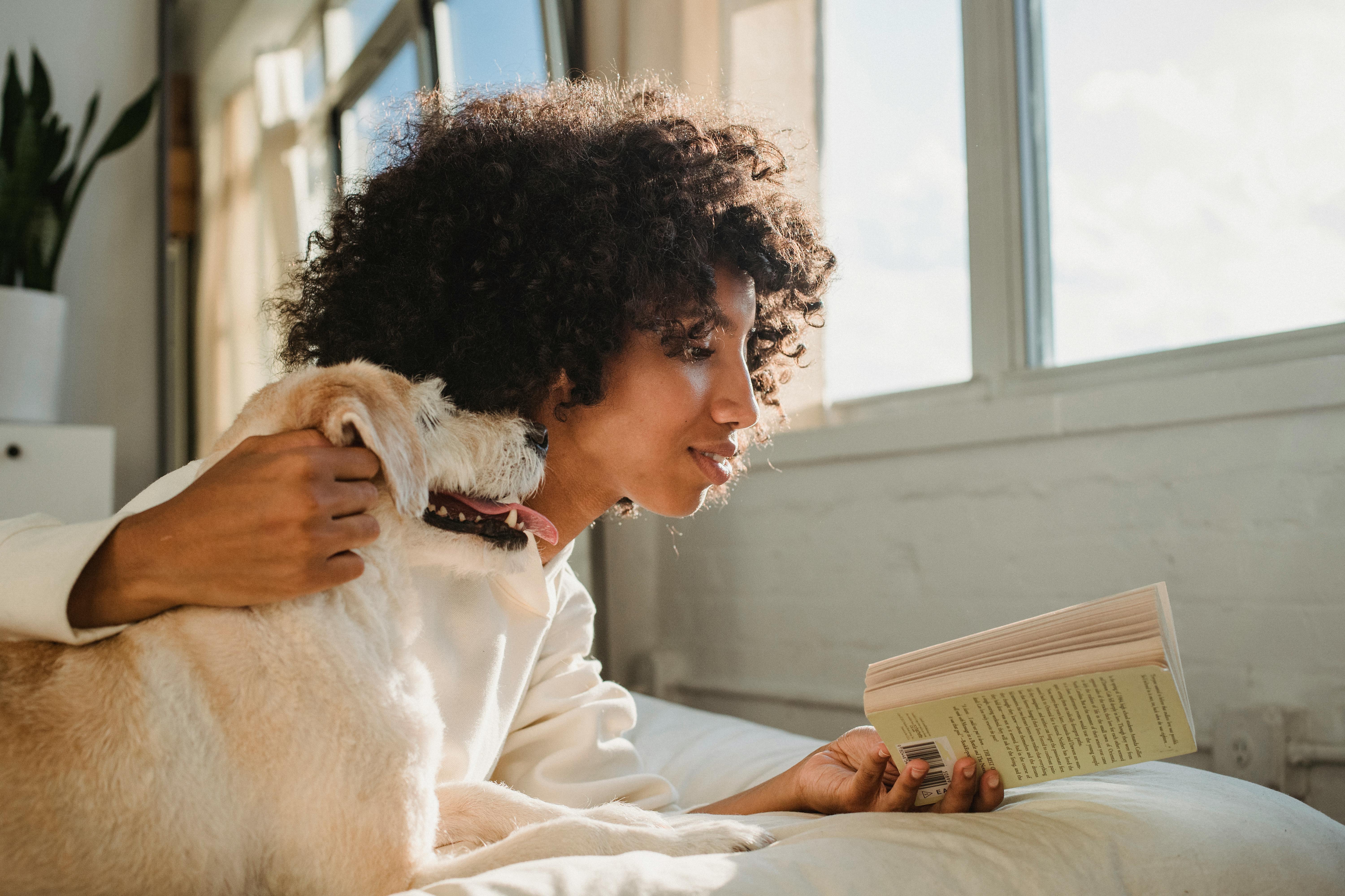 black woman caressing dog while reading book on bed