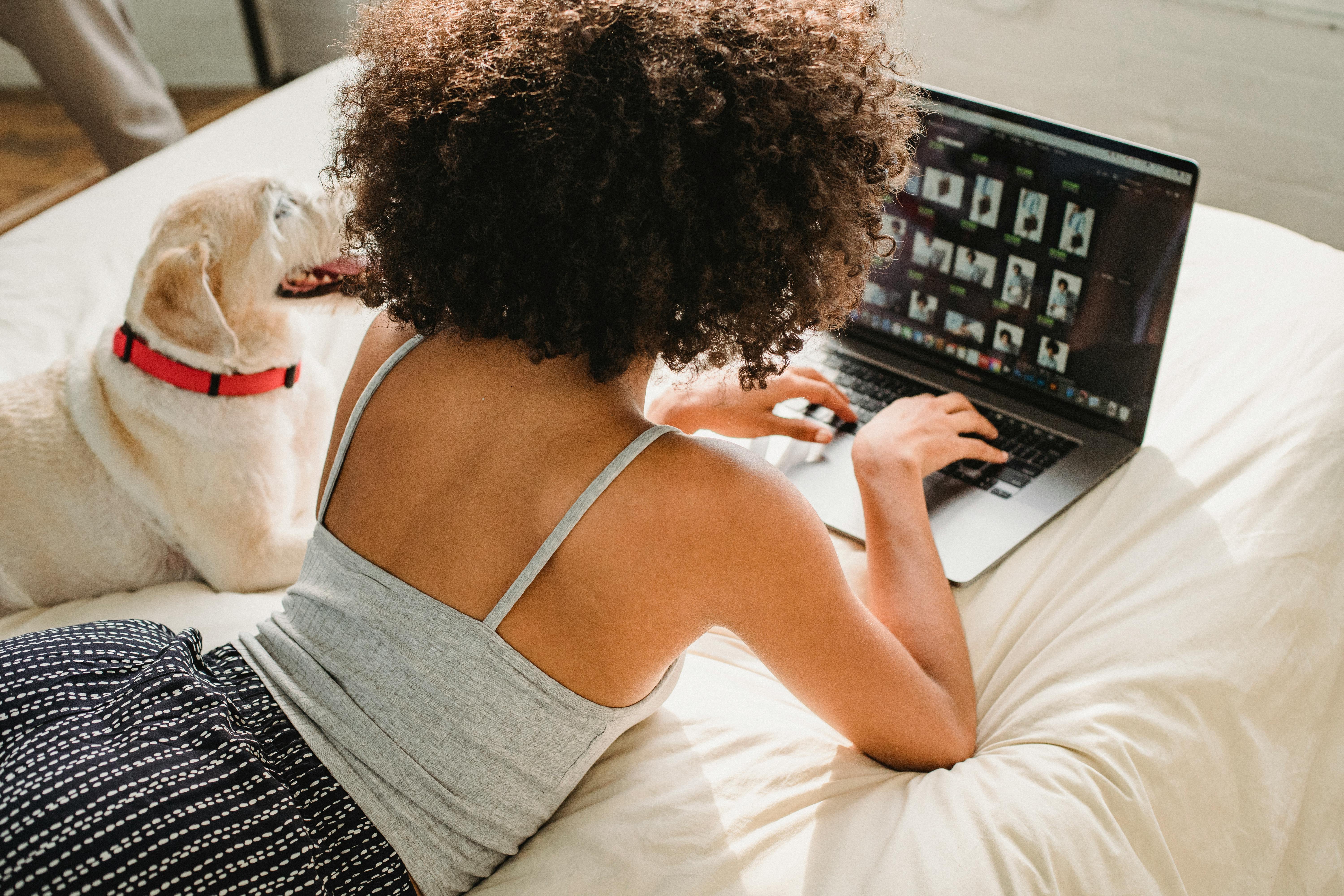 black woman browsing laptop with dog on bed