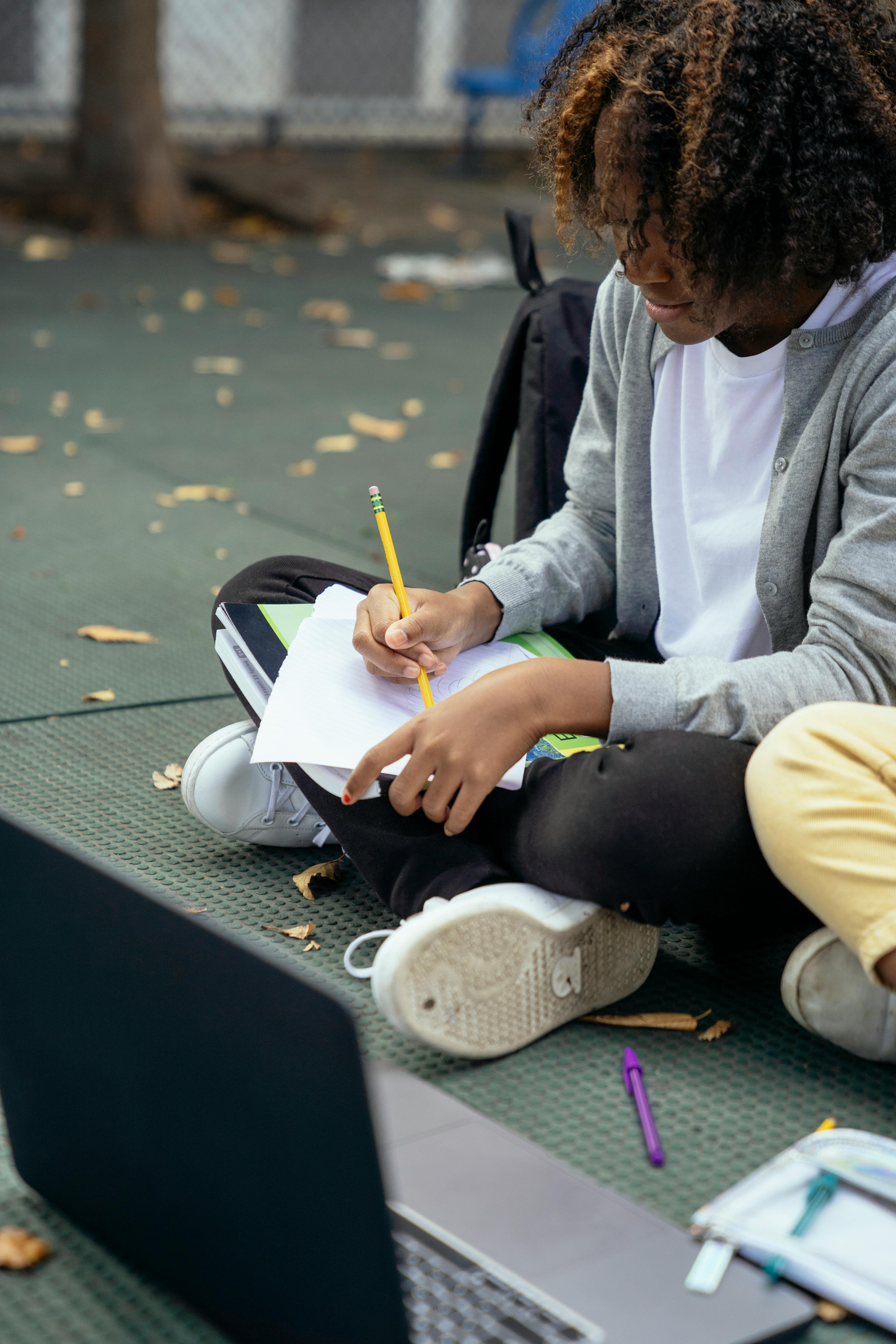 black schoolgirl writing on paper near crop friend and laptop