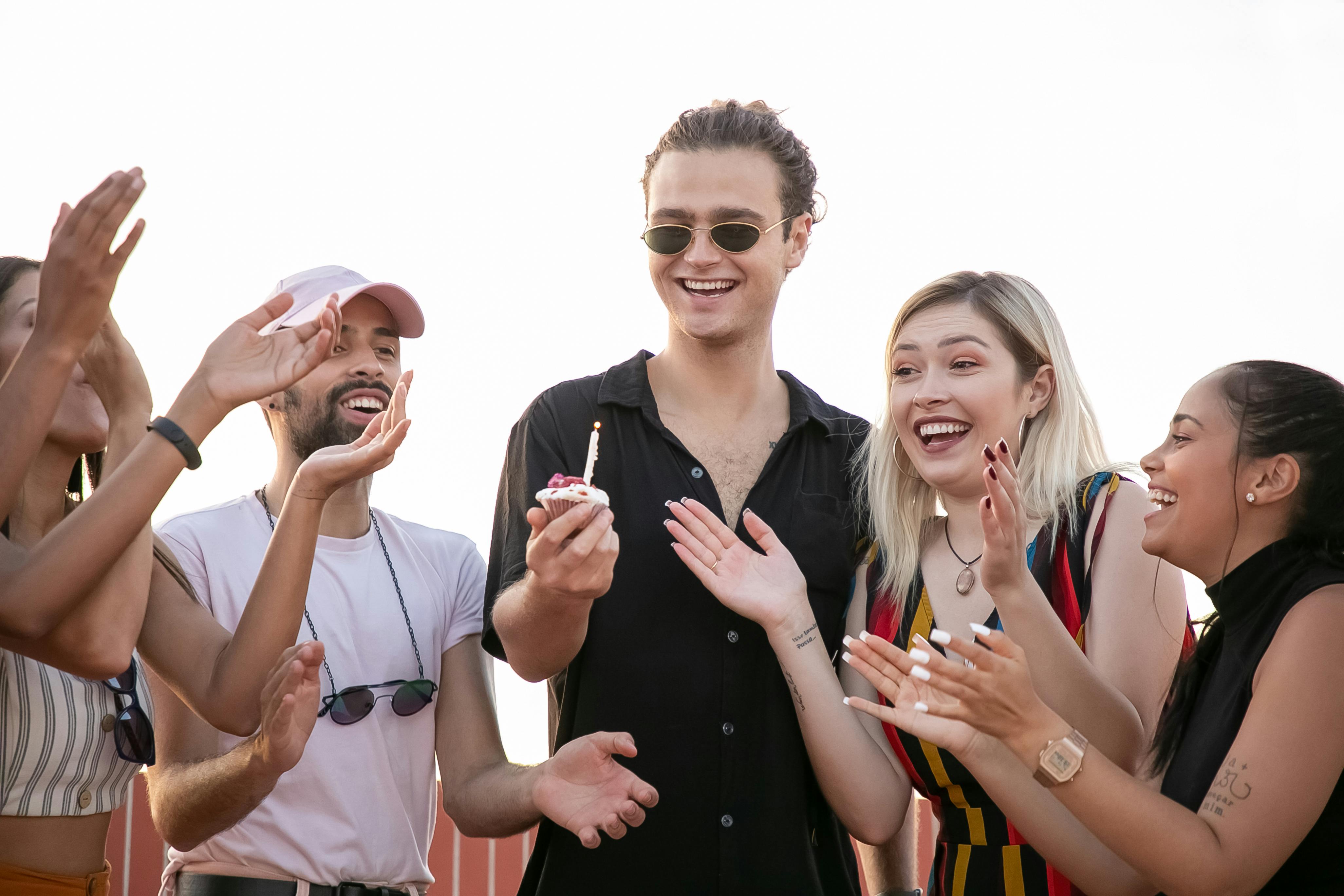 cheerful young man with birthday cupcake in hand having fun with multiracial friends