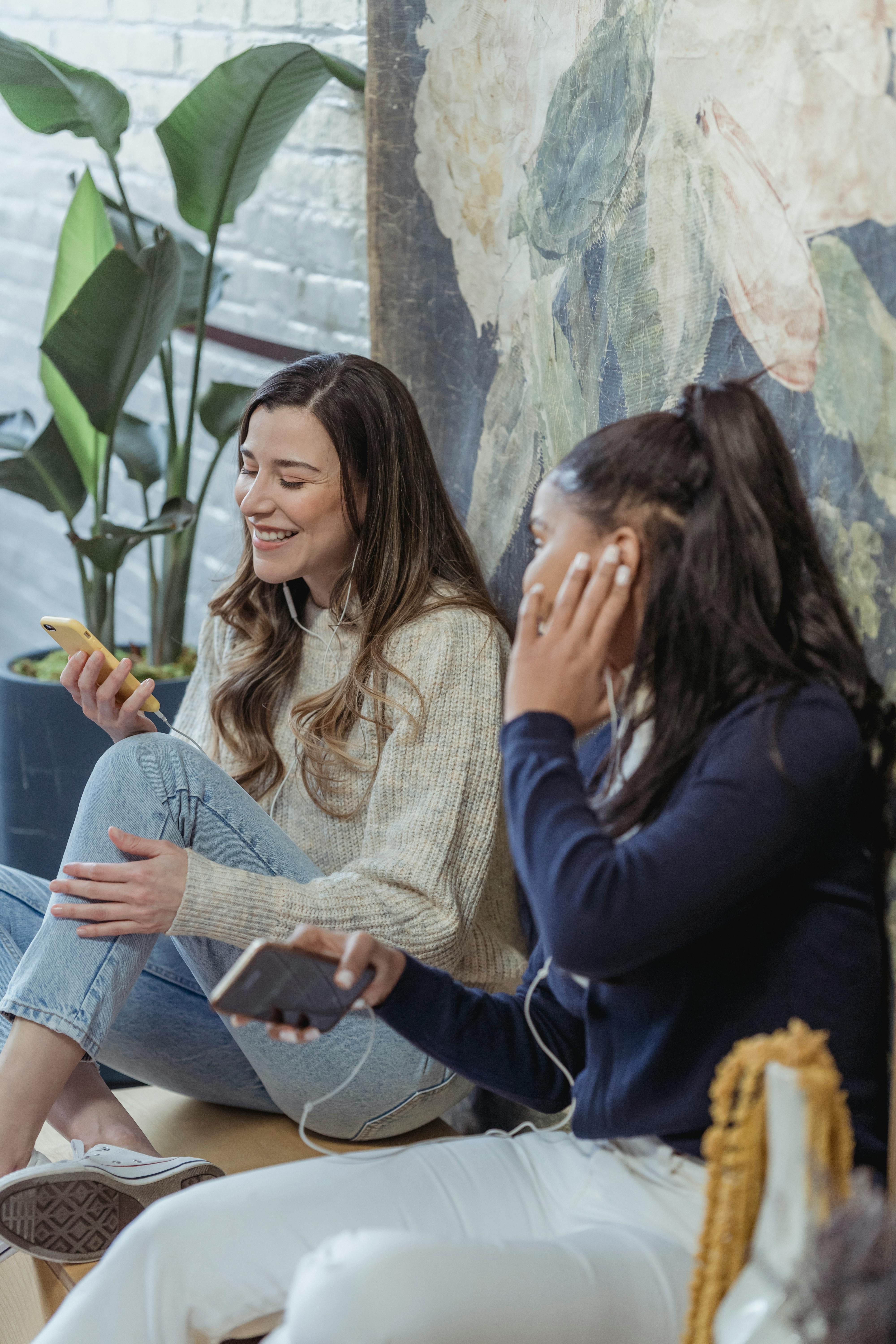 joyful young diverse ladies enjoying music in earphones while resting together at home