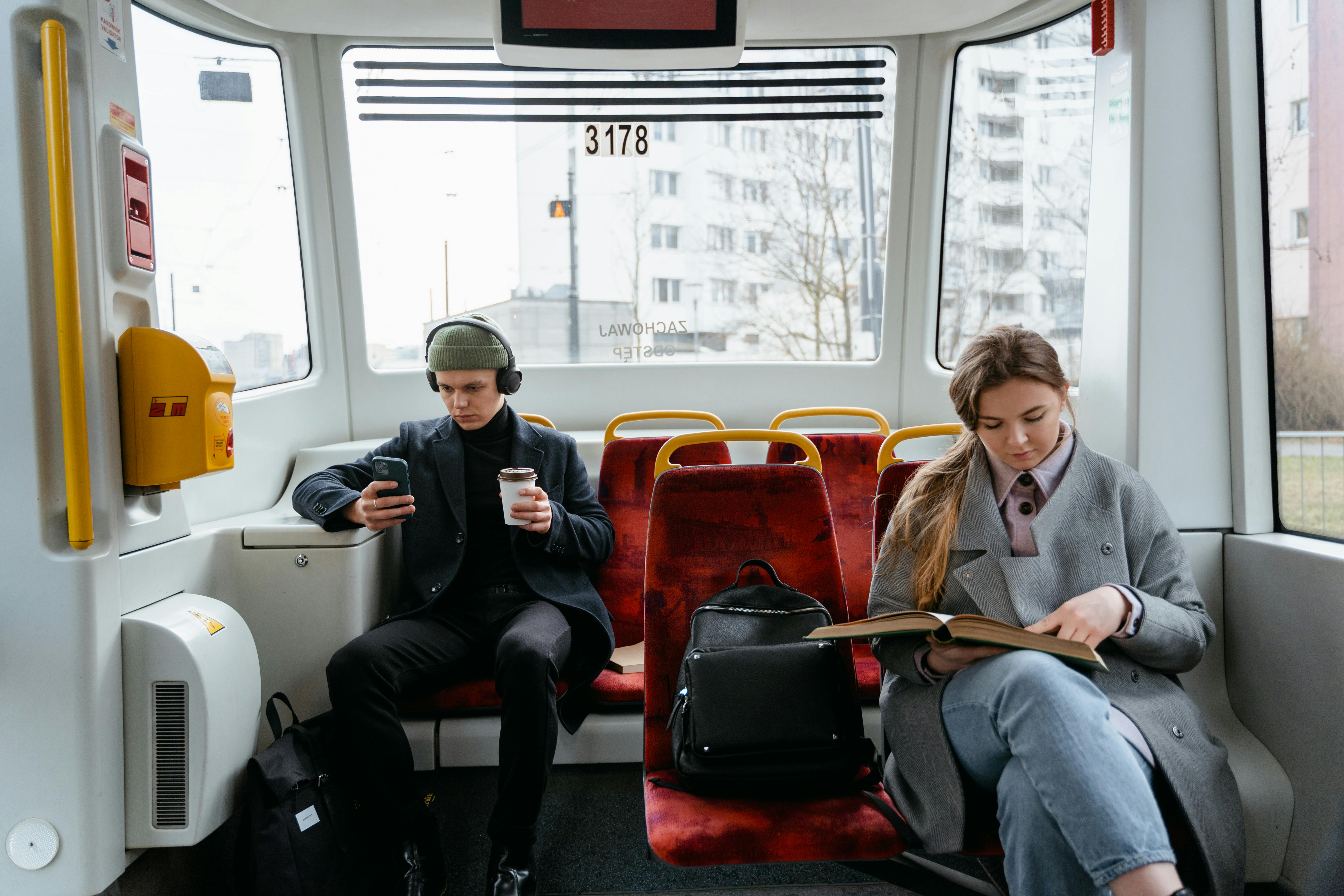 a woman in gray coat reading a book while sitting near the man wearing headphones while holding his mobile phone