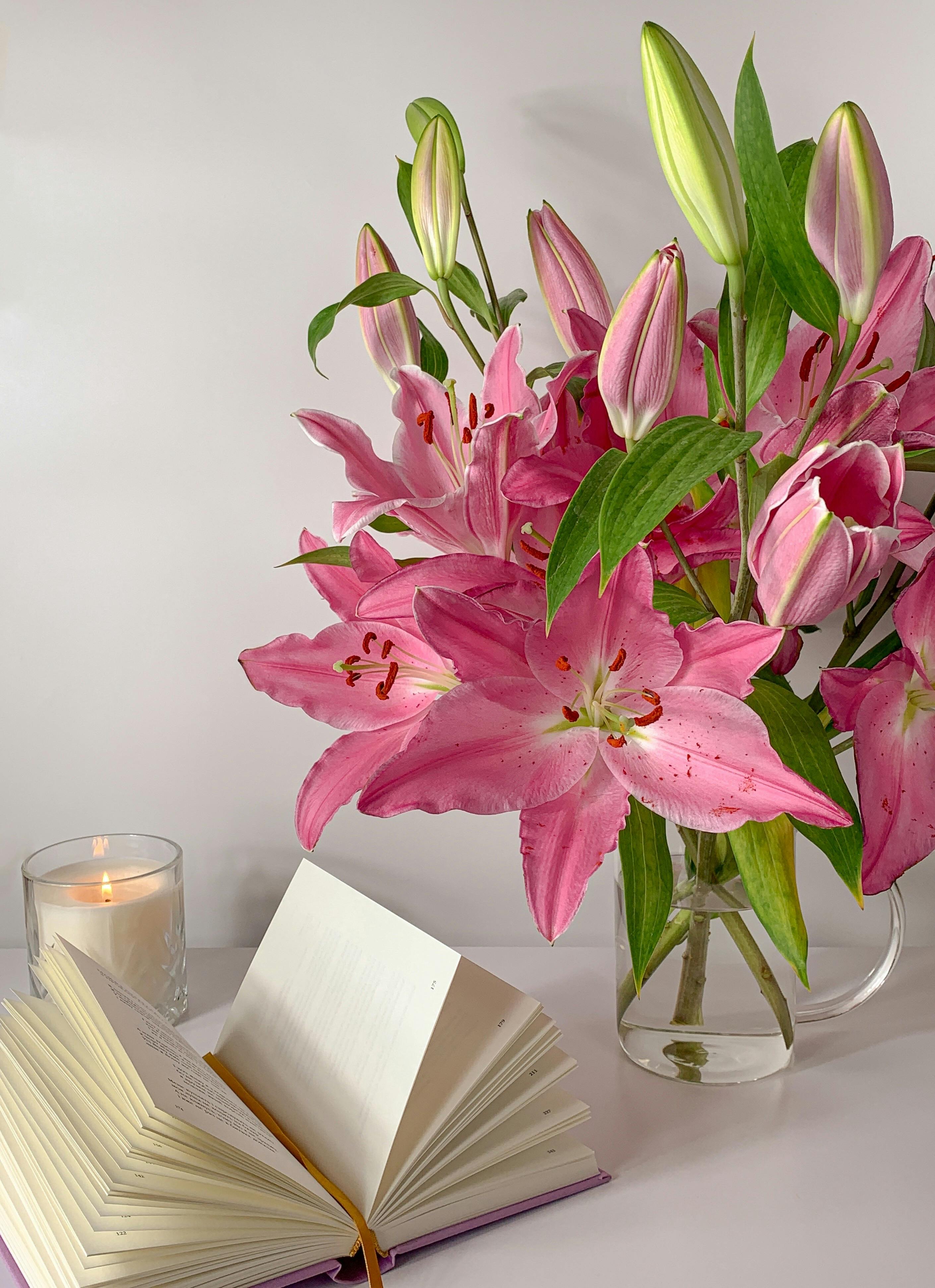 pink flowers on glass vase beside a book and lighted candle