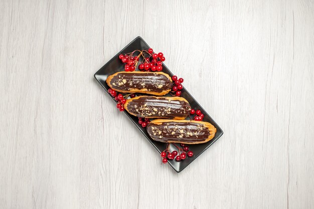 Free photo top distant view chocolate eclairs and currants on the black rectangular plate at the center of the white wooden ground