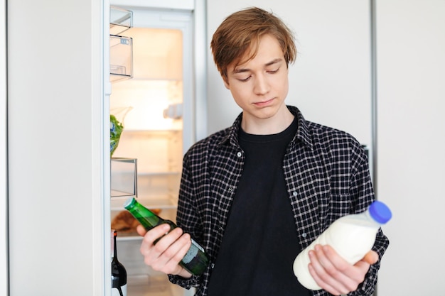 Free photo portrait of thoughtful boy standing near fridge with beer and milk in hands and deciding what to drink in kitchen at home