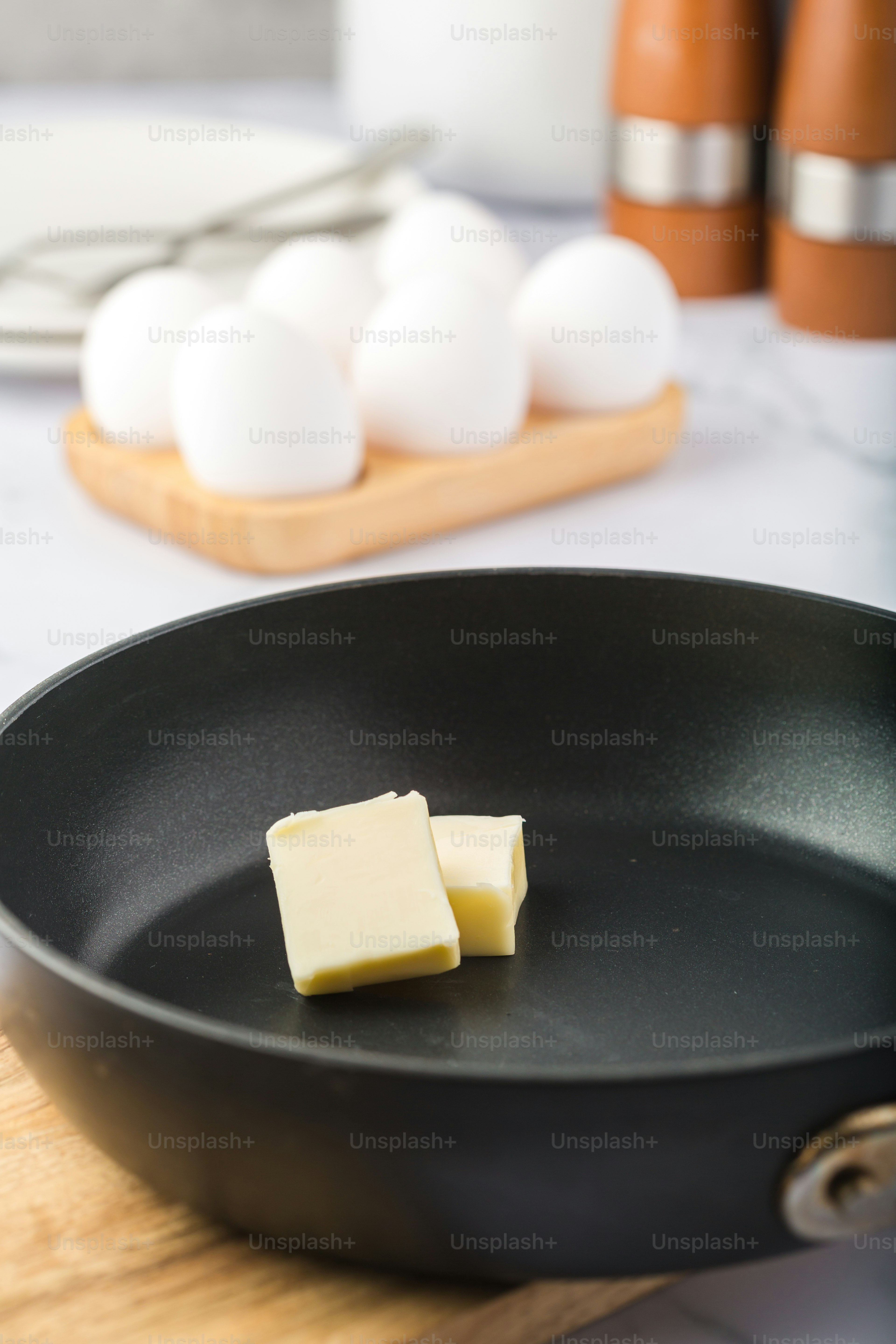 Pads of butter in a frying pan with eggs in the background.